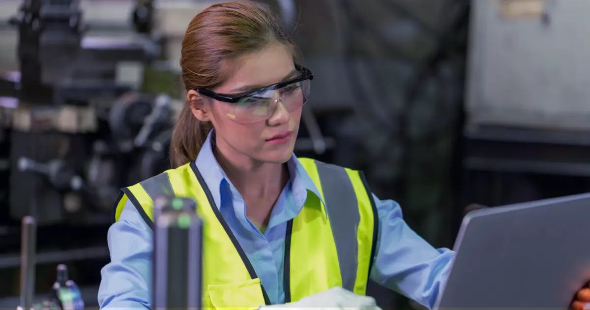 Female quality assurance engineer in safety vest using laptop at manufacturing facility, focusing on the 5 main components of Good Manufacturing Practice.